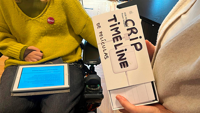 Primer plano de dos personas colaborando: una sostiene una tableta con un texto técnico sobre "Crip" y la otra sujeta una caja blanca con el título "GRIP TIMELINE DE PELÍCULAS". Se aprecia el joystick de una silla de ruedas eléctrica a la izquierda.