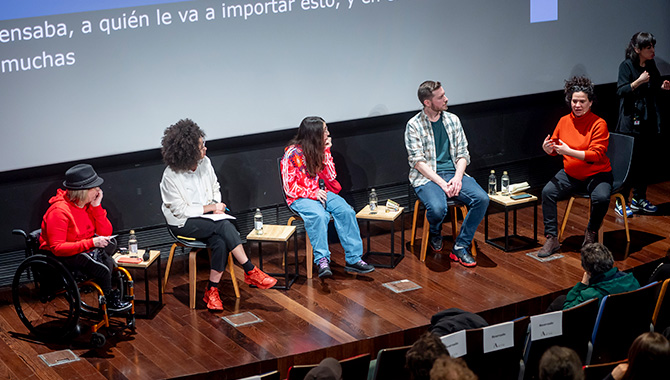 Mesa redonda en un auditorio con cinco ponentes sentados en fila y una intérprete de lengua de signos a la derecha. En la pantalla del fondo se lee un texto sobre la importancia de la participación. El público se ve desenfocado en la parte inferior.
