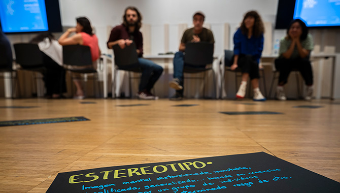 Fotografía a ras de suelo de una cartulina negra en el centro de una sala con la palabra "ESTEREOTIPO" escrita en amarillo. Al fondo, un grupo de personas sentadas en círculo conversan en una zona de taller iluminada.