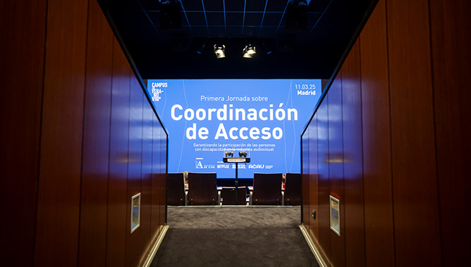 Vista desde el pasillo de entrada a una sala de cine donde una gran pantalla azul proyecta el título: "Primera Jornada sobre Coordinación de Acceso. Garantizando la participación de las personas con discapacidad en la industria audiovisual"